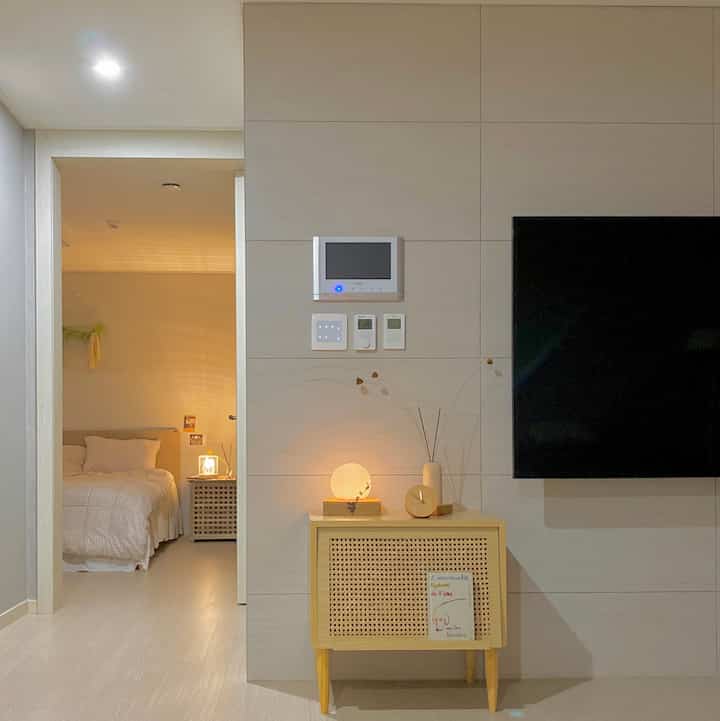 アイボリーの壁と木目調のナイトテーブルがあるミニマルなリビングルーム、覗く寝室と暖かい壁照明が調和した印象的な空間