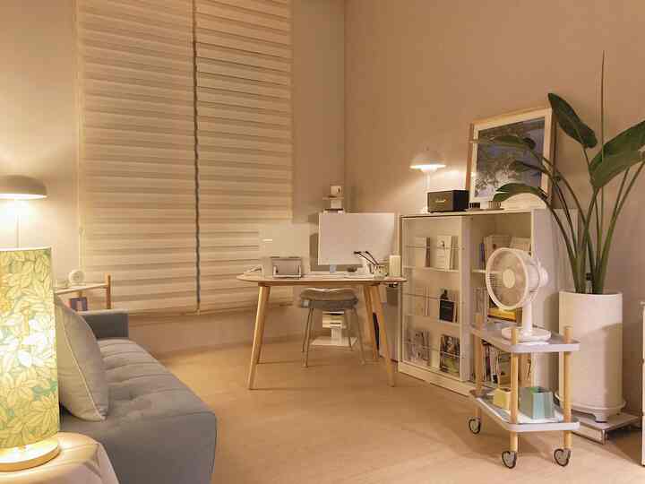 暖かいベージュトーンのホームオフィス、中央にデスクとコンピューター、左にグレーのソファ、右に収納棚が配置された雰囲気のあるインテリア