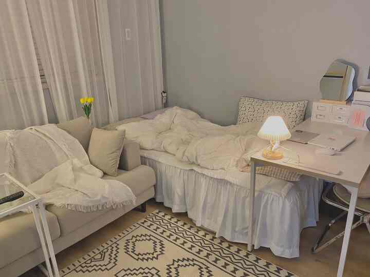 White and beige toned studio bedroom featuring sheer curtain, sofa, and desk creating cozy atmosphere