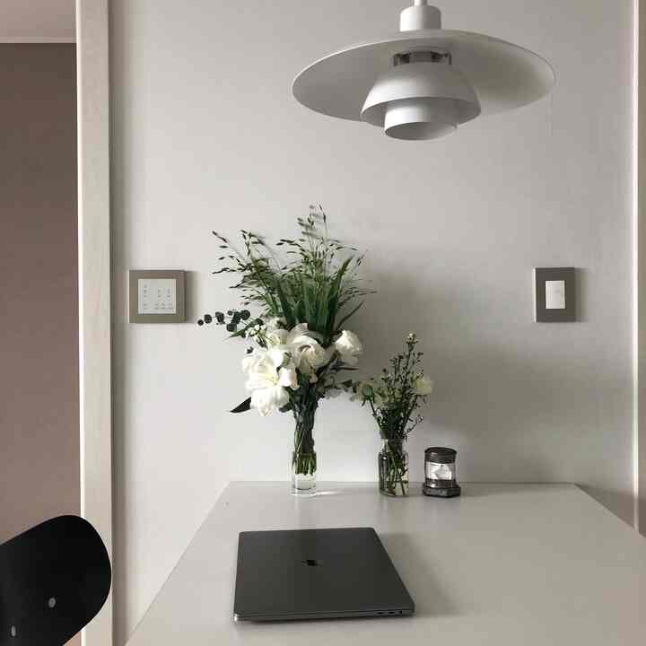 White-toned kitchen dining area featuring glass vases with flowers, laptop on table, and minimalist pendant lighting creating a clean atmosphere