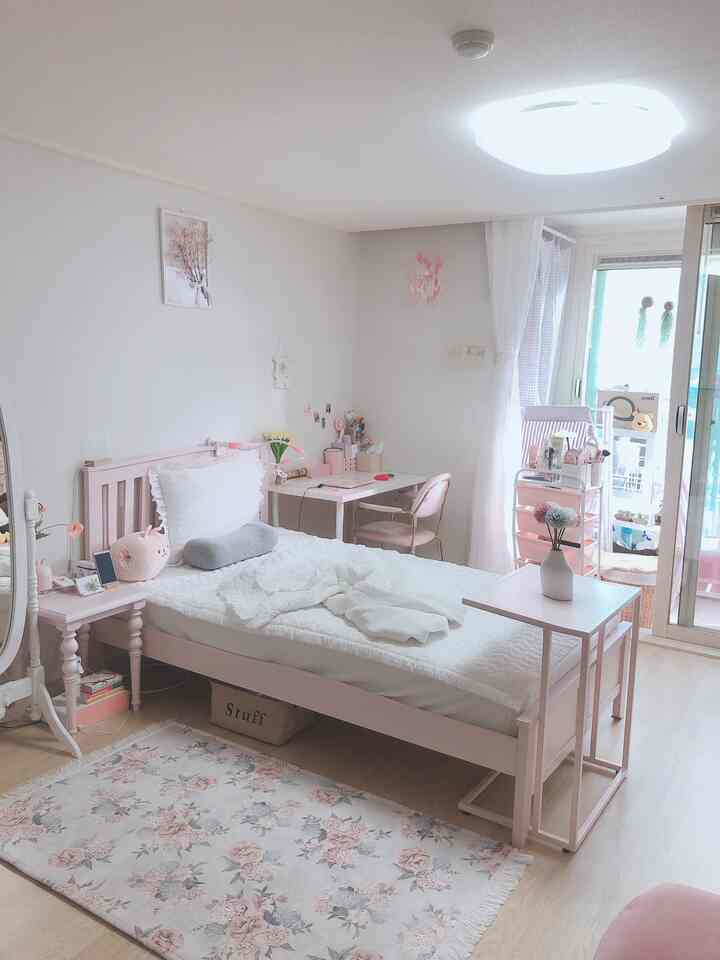 Pink and white studio bedroom featuring a pink bed, desk, balcony, and floral rug arranged cozily