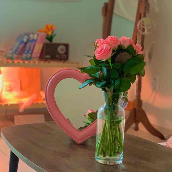 Cozy space featuring a pink heart-shaped mirror and a transparent glass vase with pink roses on a wood-toned table