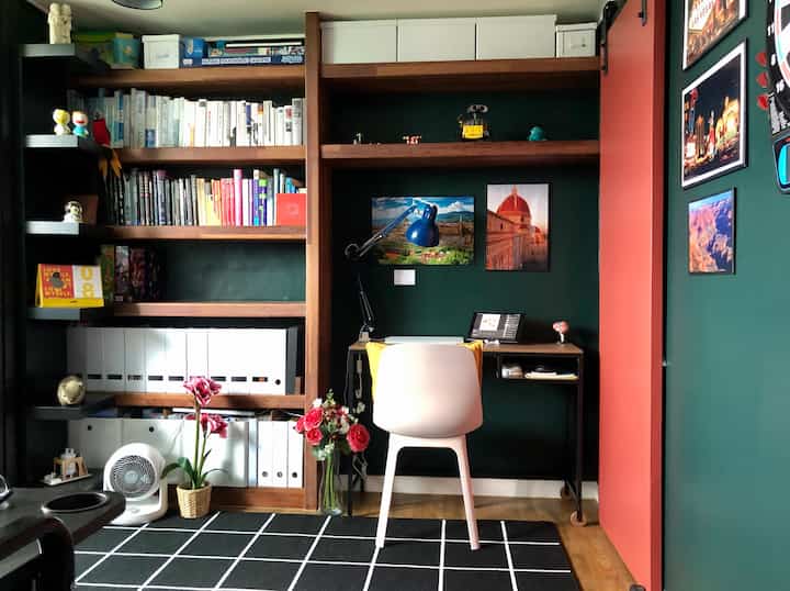A study with dark green walls and wood tones, featuring a bookshelf on the left and a desk with a white chair centrally placed, neat workspace