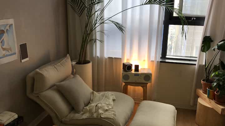 Beige and ivory toned living room featuring a high-back single recliner sofa and ottoman, with audio system on a stool by the window creating a warm relaxation atmosphere