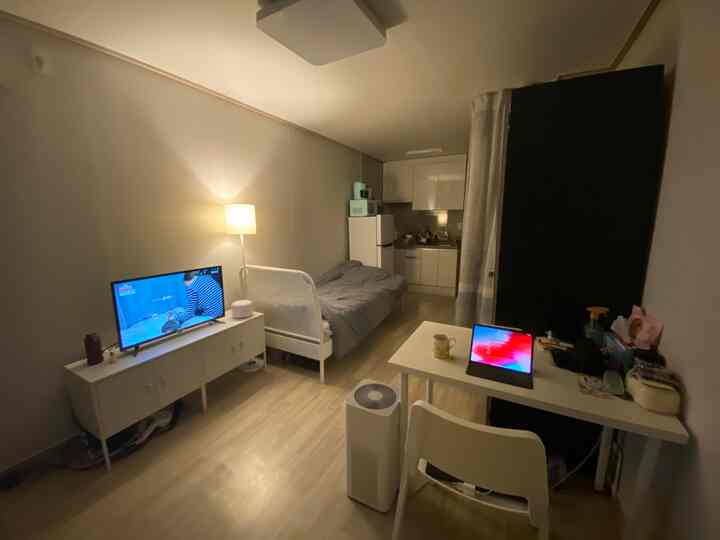 White and brown toned studio apartment bedroom and kitchen space featuring a desk and bed in a cozy single living room