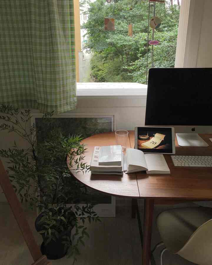 Wood tone round dining table and white window with green plants and green check curtains in a cozy home office space