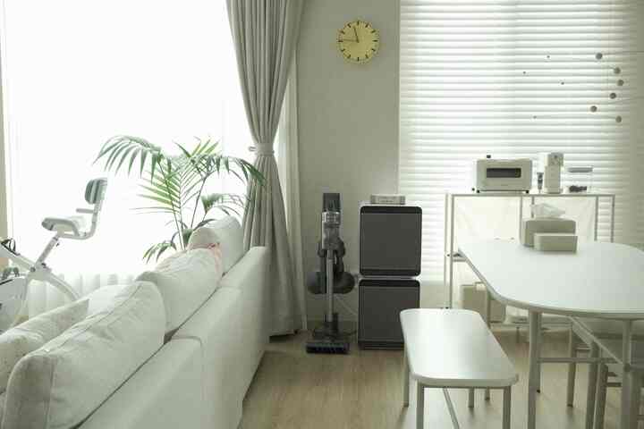 明るくホワイトトーンとナチュラルな木目の床が調和したリビング、ホワイトのモジュラーソファとダイニングテーブル、多彩な家電と植物が配置された清潔感のある空間