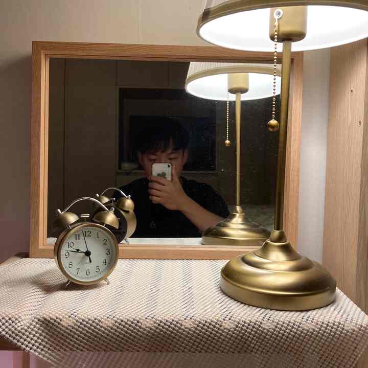 Wood-framed mirror with a gold table lamp and vintage clock on a textured surface, evoking a cozy bedroom setting
