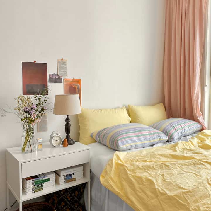 柔らかな黄色と白の色調の寝室、黄色い寝具と白いナイトテーブルにテーブルランプと花瓶が配置されたシンプルでナチュラルなインテリア
