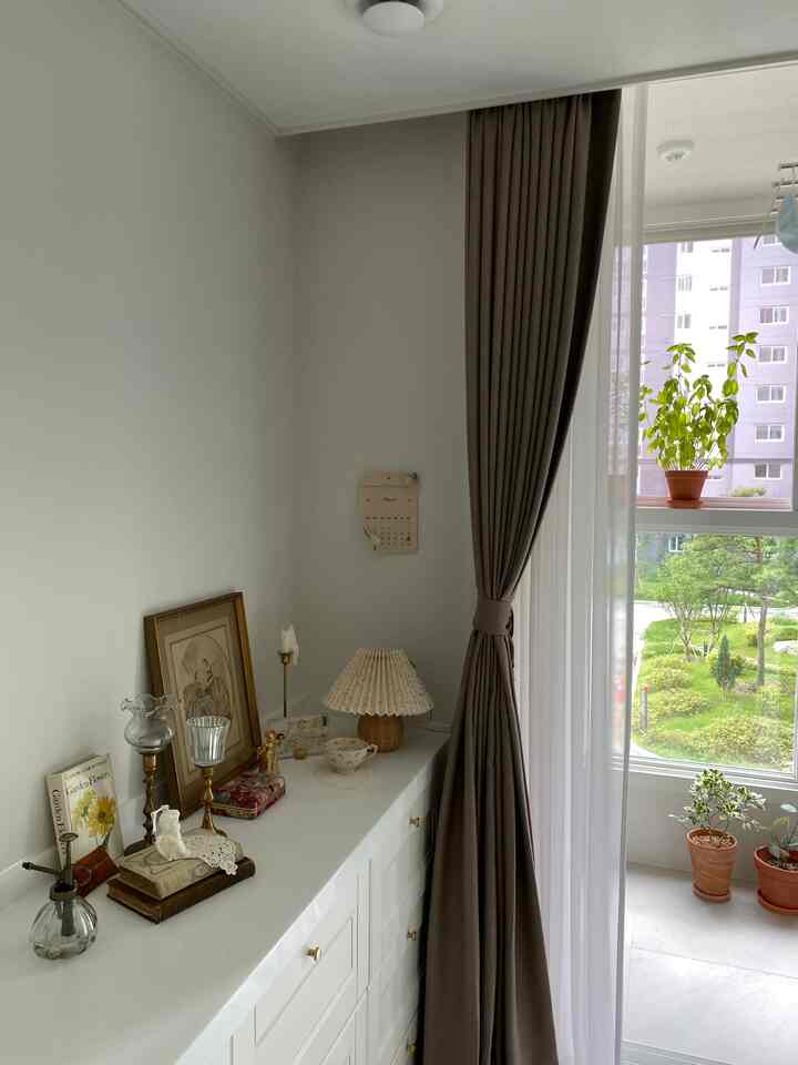 Beige blackout curtain and white built-in dresser by a windowed bedroom featuring vintage decor and plants in a cozy healing zone