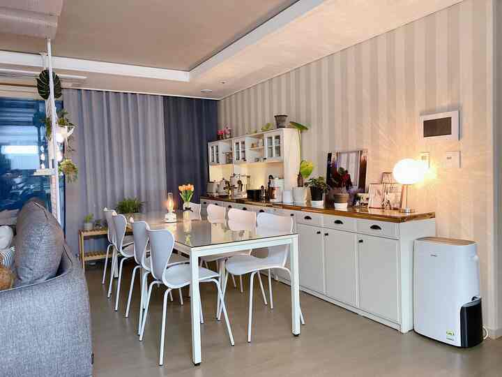 White and navy toned living and kitchen space featuring an 8-person dining table, various coffee machines, plants, and warm lighting with a cozy home cafe atmosphere
