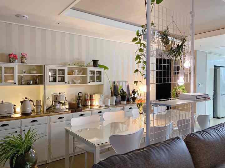 White and brown toned living room with a home cafe setup, featuring a dining table, grid room divider, plants, and coffee machine creating a cozy atmosphere