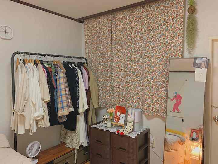 ベージュと木目調のワンルーム、クローゼット用ハンガーラック、花柄カーテン、全身鏡、収納棚が配置された雰囲気のあるインテリア