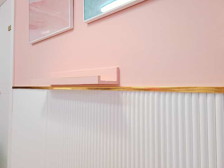Modern interior space featuring pink upper wall, white lower panels, gold accent shelf, and picture frames in harmony