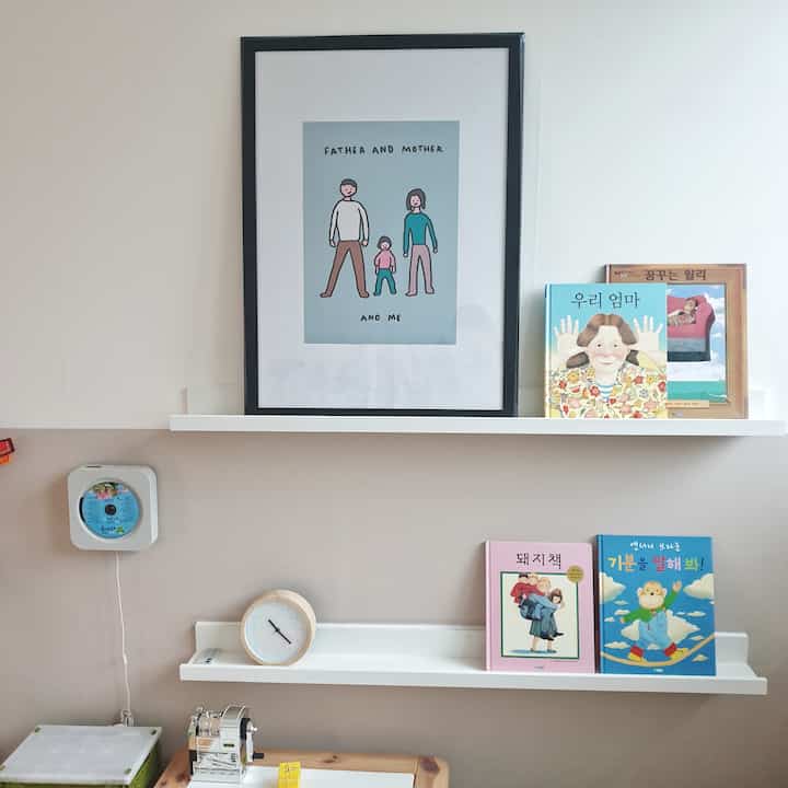 クリーム色とトーンダウンピンクの壁の子供部屋、壁の棚には絵画や子供の本が置かれた雰囲気のあるインテリア