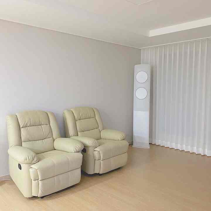 明るいベージュ調のリビング、2台の一人用リクライニングソファと白いエアコン、ナビプリーツカーテンが配置されたミニマルな空間