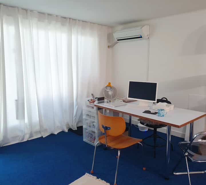 白い壁とカーテンに青いカーペットが敷かれたコンパクトなホームオフィス、机にコンピュータとオフィスチェアが配置された整った空間