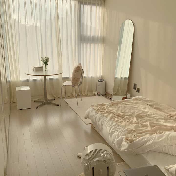 Creamy ivory toned studio bedroom featuring a bed, round table, and full-length mirror with a cozy natural atmosphere