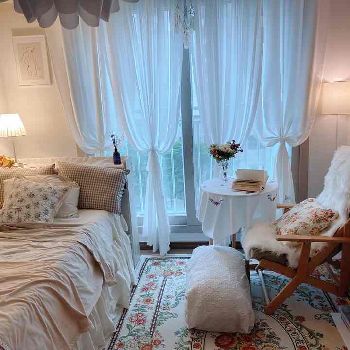 Bright studio apartment in white and natural tones featuring armchair, Persian rug, and sheer curtains creating cozy atmosphere