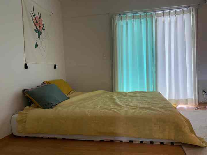 明るい白い壁とカーテンがある6畳の寝室、黄色いベッドカバーと緑のクッションが安らぎのあるインテリア