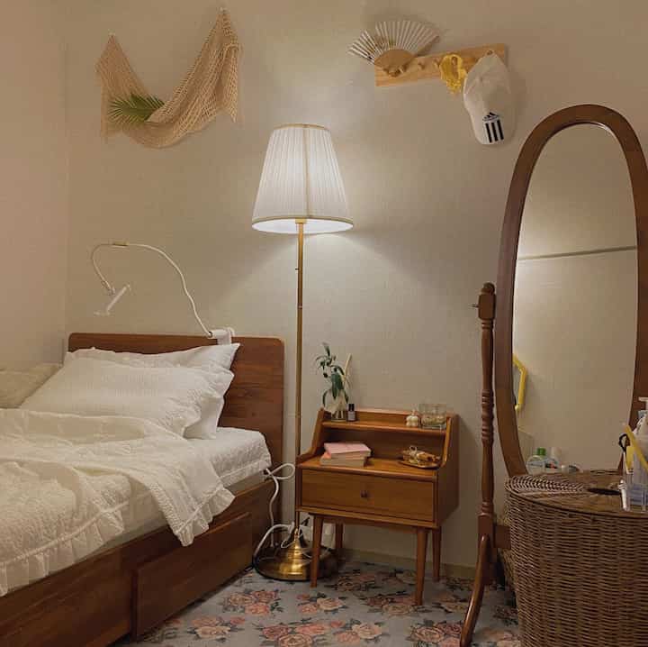 Natural wood and vintage styled small bedroom featuring a wooden bed, side table, floor lamp, and full-length mirror for a cozy atmosphere