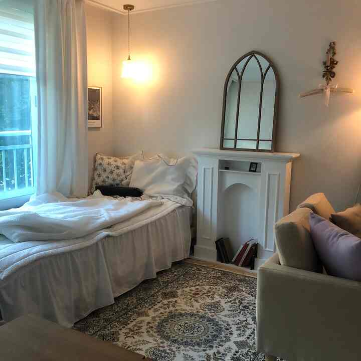 White and natural toned bedroom featuring a vintage mirror above a fireplace console, cozy sofa, and patterned rug creating a warm atmosphere