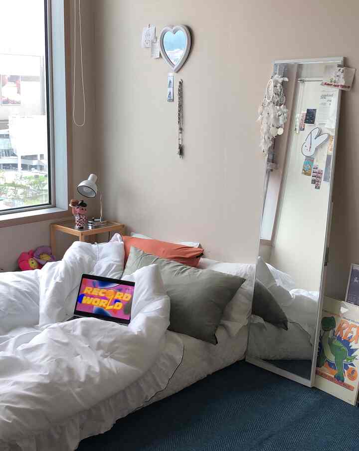 ベージュ色の壁と白い寝具が配置されたワンルームの寝室、オリーブグリーンとオレンジのクッション、木目調のサイドテーブルが調和した居心地の良い空間