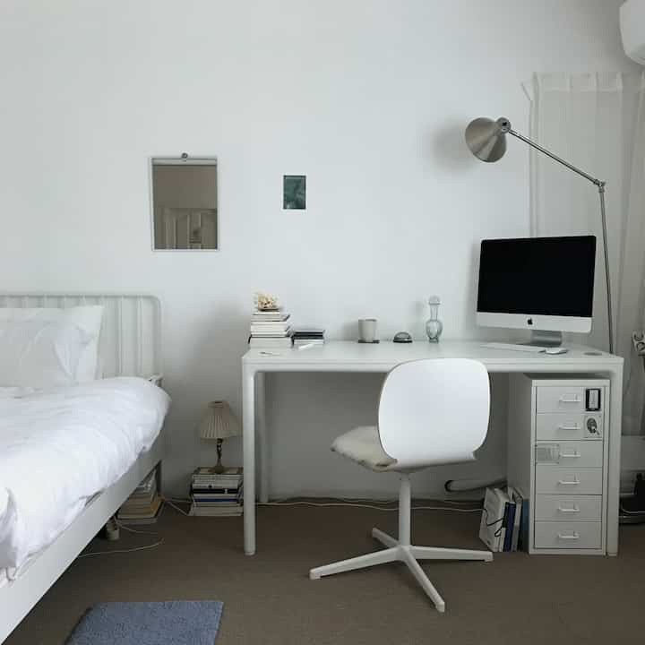 白を基調とした小さな寝室、ベッド横にデスクを置き、本やランプが並べられたシンプルなホームオフィス空間