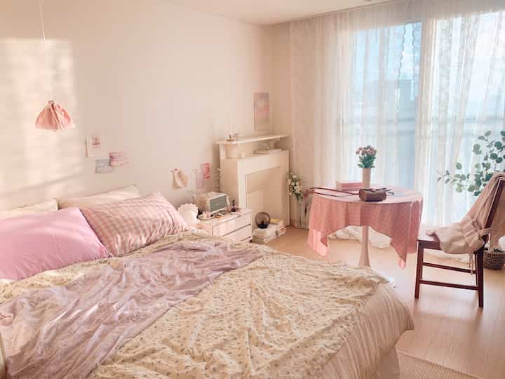 A bedroom featuring pink and wood tones, lace curtains, and a small table creating a warm and cozy atmosphere