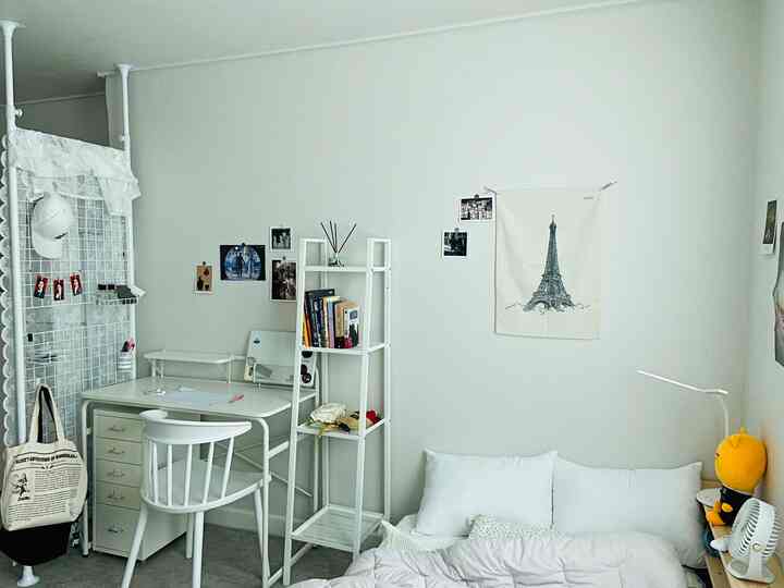 White-toned bedroom featuring a desk, shelf, bed, and posters creating a simple, cozy atmosphere
