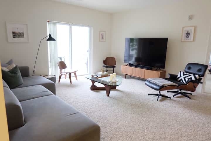 Bright white-toned living room with carpet flooring featuring gray sofa, black leather lounge chair, and wood-glass coffee table in a Mid-Century Modern style