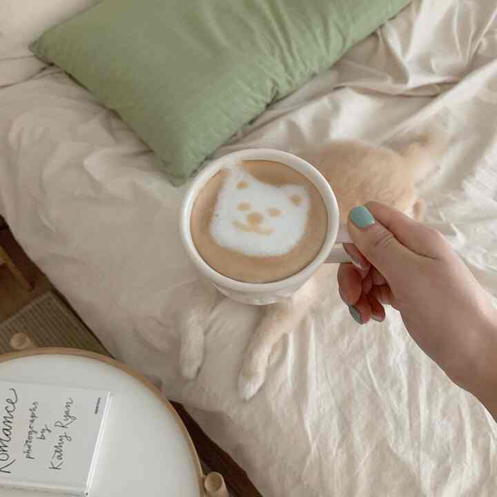 ベージュのナチュラルカラーの寝室、ベッドに犬が横たわり、手に犬のラテアートのマグがある雰囲気のあるインテリア