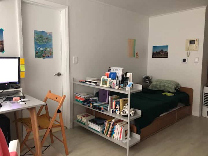 A small, cozy single household bedroom with white walls, wood-tone flooring, featuring a desk and bookshelf, practical setup