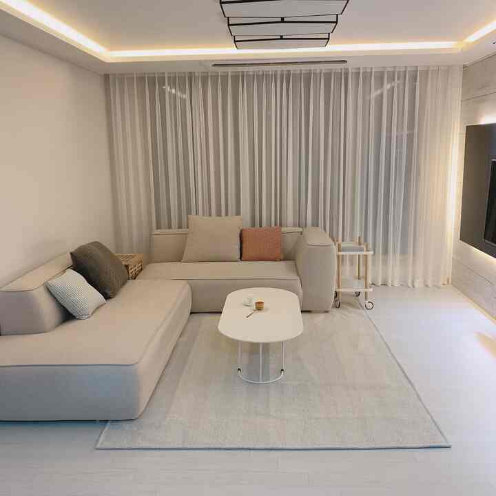 A white and beige toned living room featuring a large modern sofa, white curtains, and a light gray rug creating a cozy atmosphere