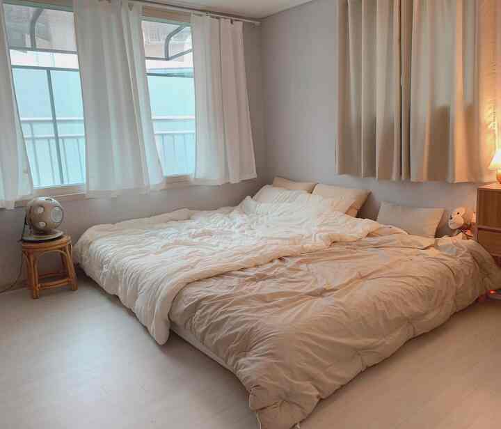 Beige and white toned bedroom featuring a low bed, blackout curtains, and a rattan stool creating a calm atmosphere