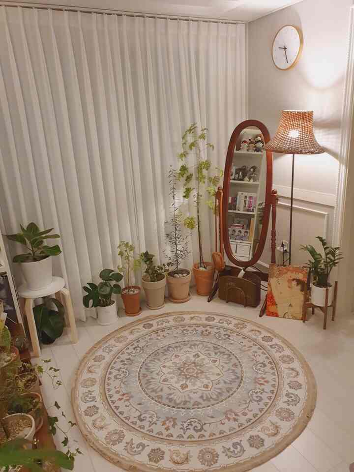 A white and wood-tone living room featuring various indoor plants, a wooden full-length mirror, round rug, and rattan floor lamp creating a cozy atmosphere