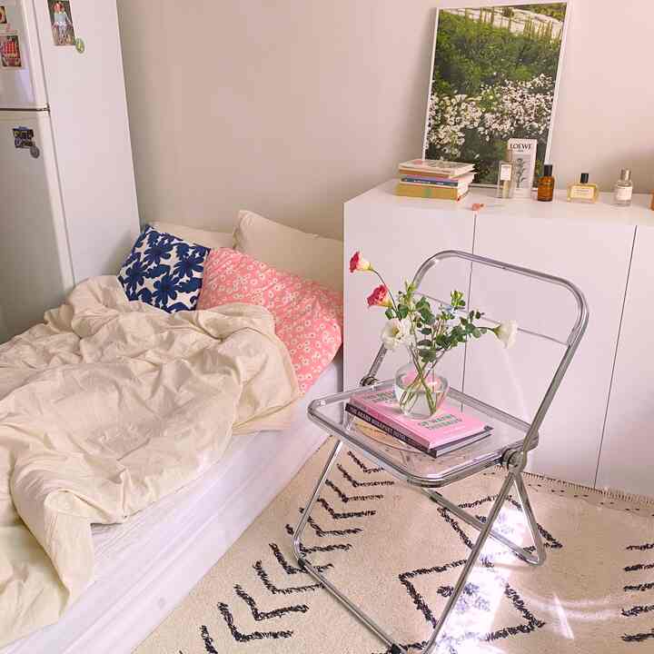 白い壁とベージュの寝具があるワンルーム寝室、透明な折りたたみ椅子に花瓶と花が置かれ、リズミカルなラグがある雰囲気のよい空間