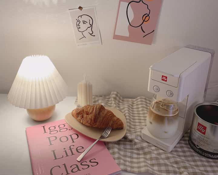White and beige toned home cafe featuring coffee machine, croissant, and table lamp creating a cozy atmosphere