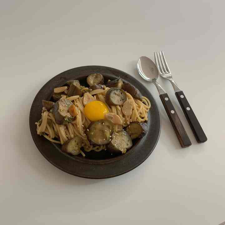 Dark brown plate with cutlery on white table featuring eggplant pasta dish