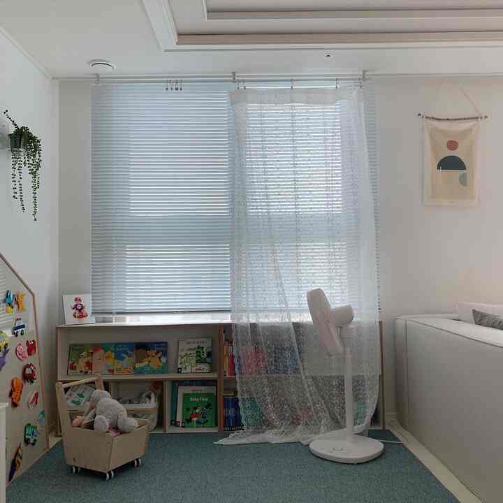 Bright natural-tone living room with white blinds and lace curtains by the window, cozy kids' bookshelf and toys arranged in play area