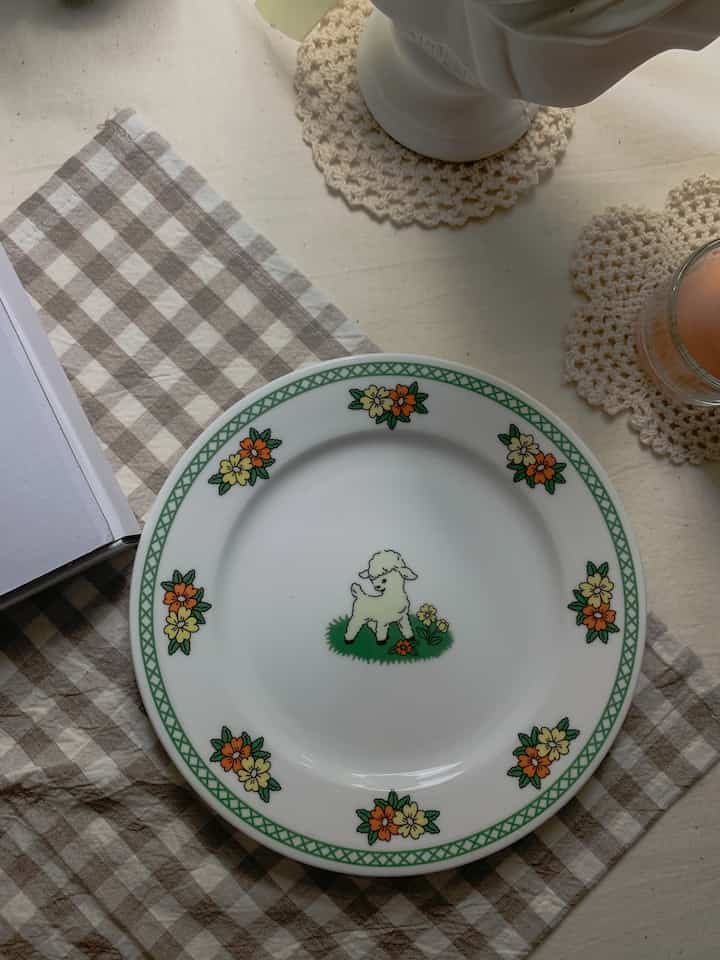 Beige checkered placemat with a plate featuring a lamb and floral pattern and crocheted coasters creating a cozy table setting
