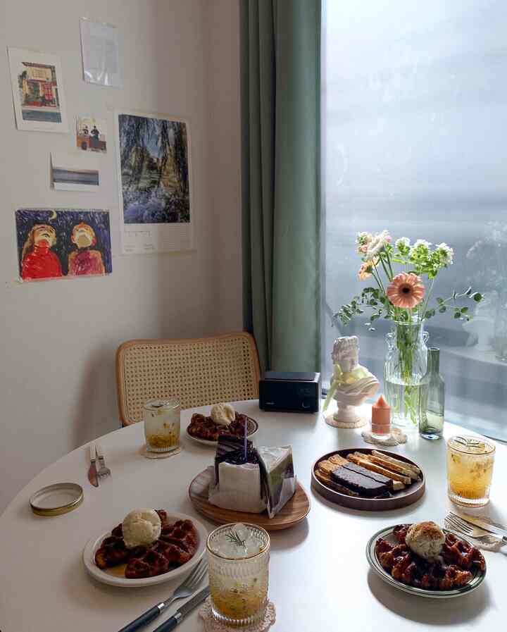A modern, natural home cafe dining room with a dominant white table, wood-toned chair, green curtains, and flower decorations