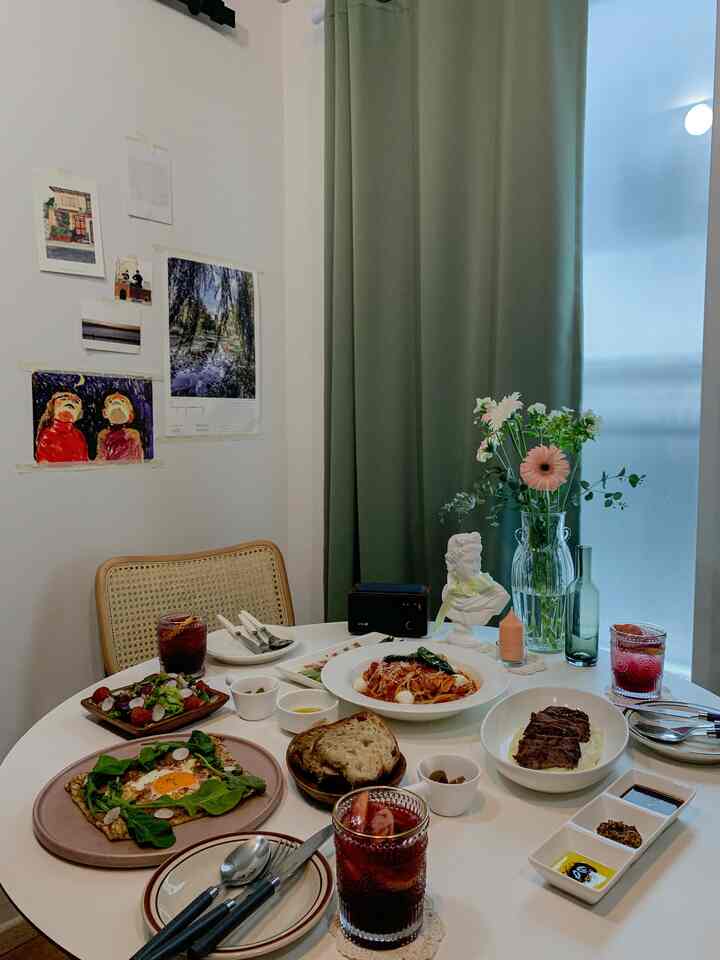 白い壁と丸いダイニングテーブル、緑の遮光カーテンがあるアットホームなダイニングルーム、花瓶と多彩な料理が並ぶ空間