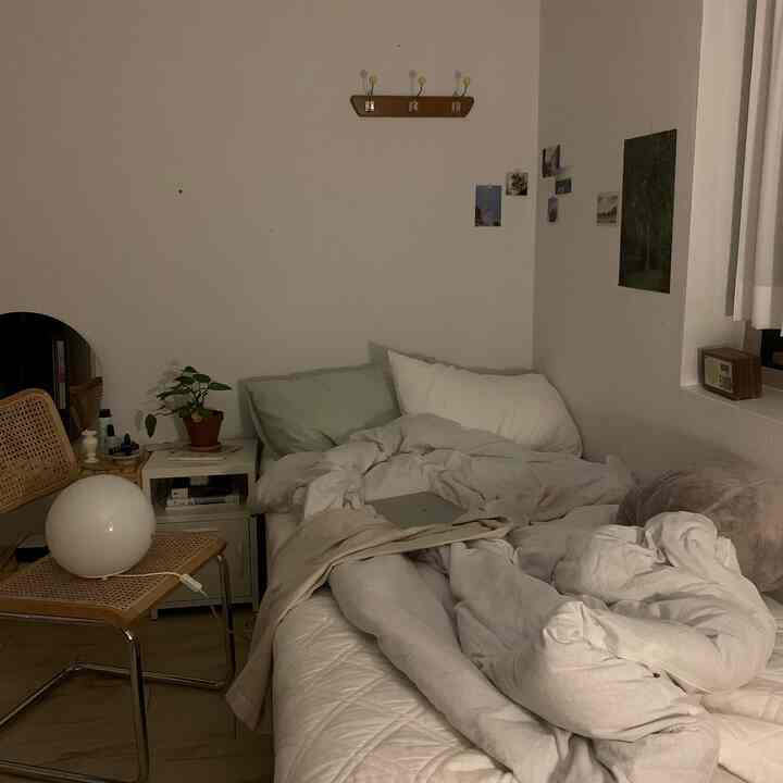 Simple studio bedroom with white and natural tones featuring bed, nightstand, rattan chair, and plant