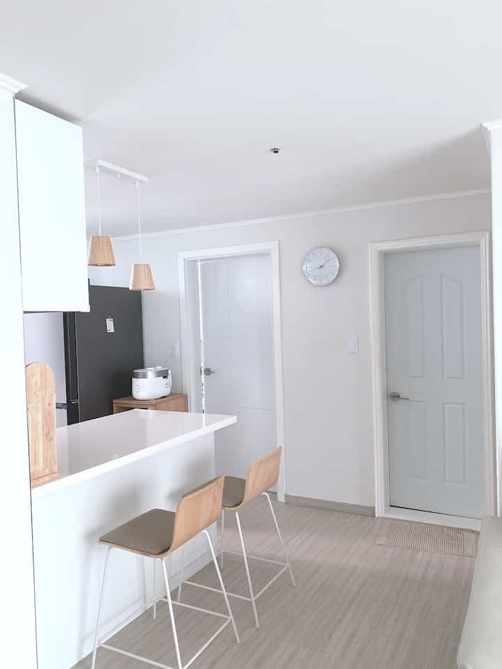 白と木目調が調和するキッチン、キッチンアイランドと木製ペンダントライト、バースツールが配置されたシンプルで明るい雰囲気のインテリア