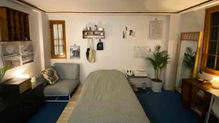 ナチュラルな木目調とブルーの床が調和した一人暮らしの寝室で、中央にベッド、左にソファ、右に全身鏡と植物が配置された雰囲気のあるインテリア