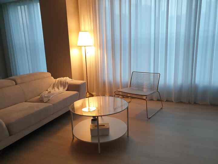 A simple modern living room with white and wood tones featuring a sofa, coffee table, metal chair, and soft floor lamp lighting