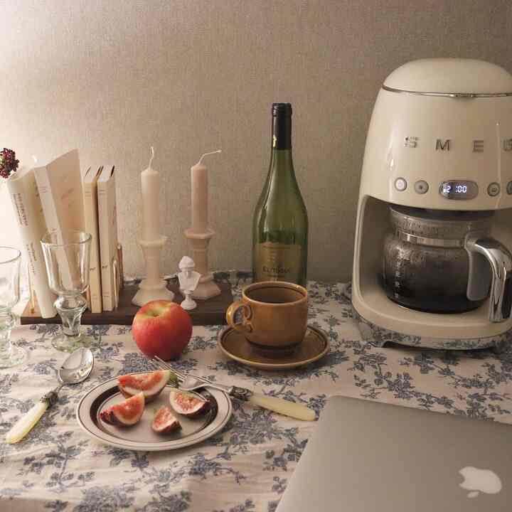 Natural beige toned home cafe space with floral tablecloth, featuring coffee machine, candles, books, and fruit creating a cozy atmosphere