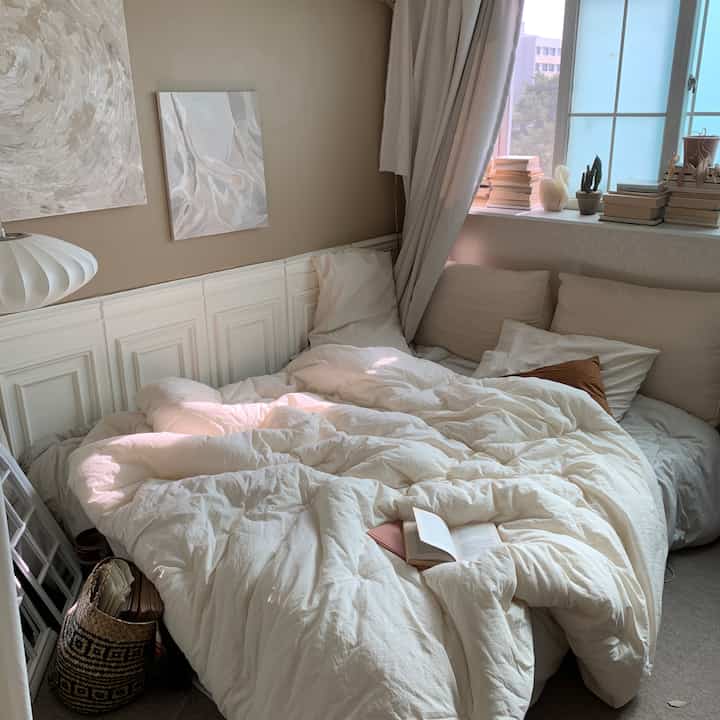 White and beige toned bedroom featuring white bedding, curtains, and books on the windowsill creating a cozy, neat atmosphere
