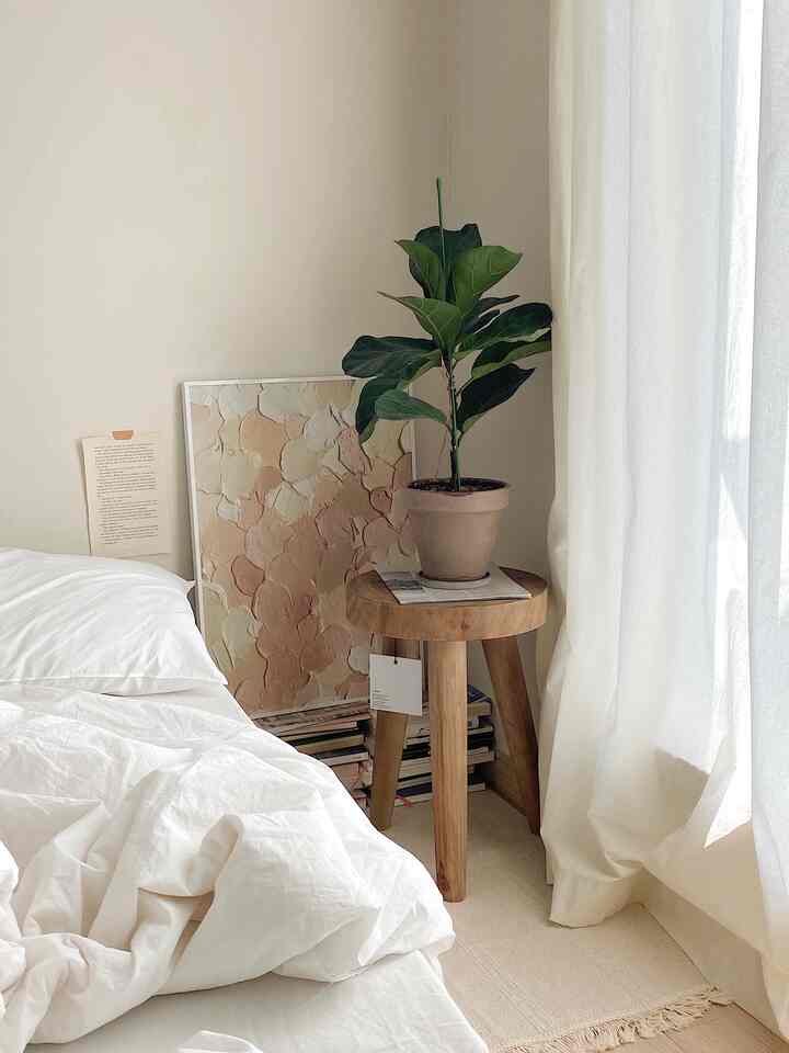 Beige wall and white bedding in bedroom featuring wooden stool with potted plant and poster, natural modern style